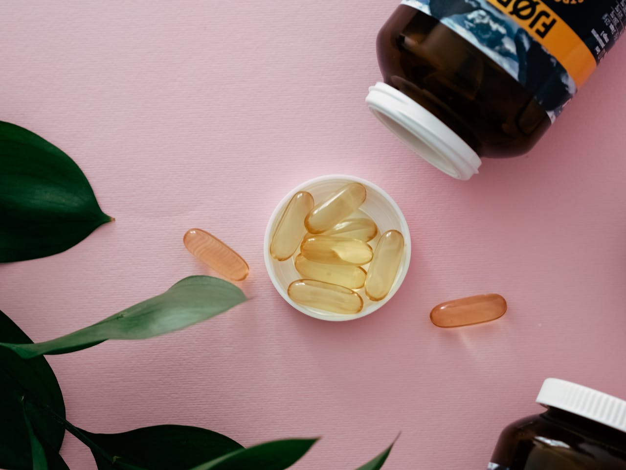 Top view of health supplements with bottles on a pink background, featuring capsules and green leaves.